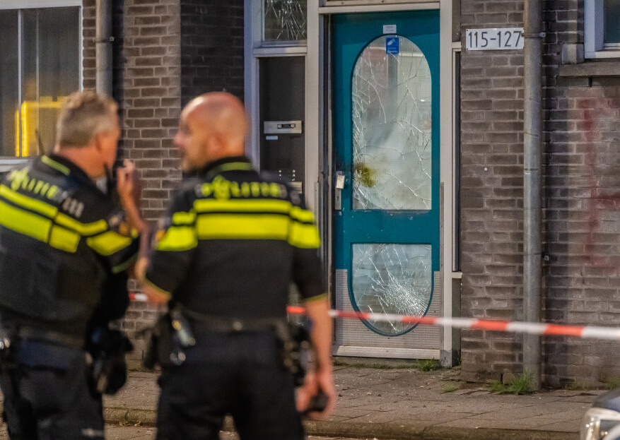 twee agenten staan achter afzetlint bij een deur met gebroken ramen